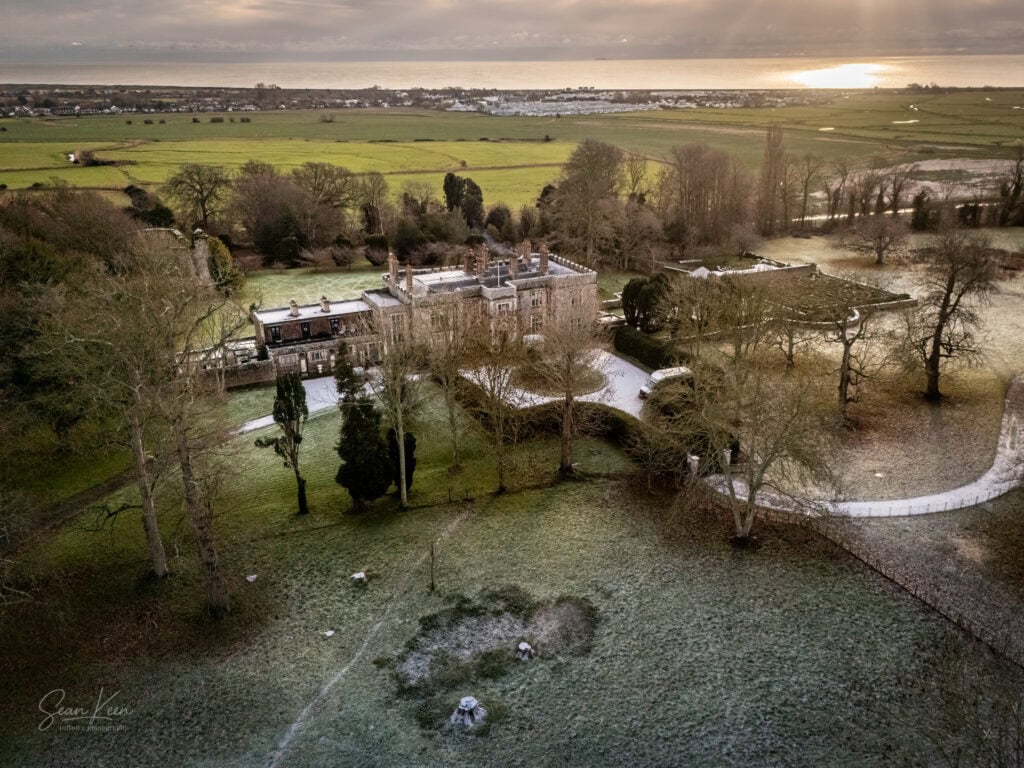 Aerial view of a large historic stone mansion, ideal for wedding photography, surrounded by trees and frosty grass, with winding paths and open fields stretching toward a coastal horizon under a cloudy sky reflecting sunlight on the sea. Infinity Photography - Wedding Photographer - East Sussex