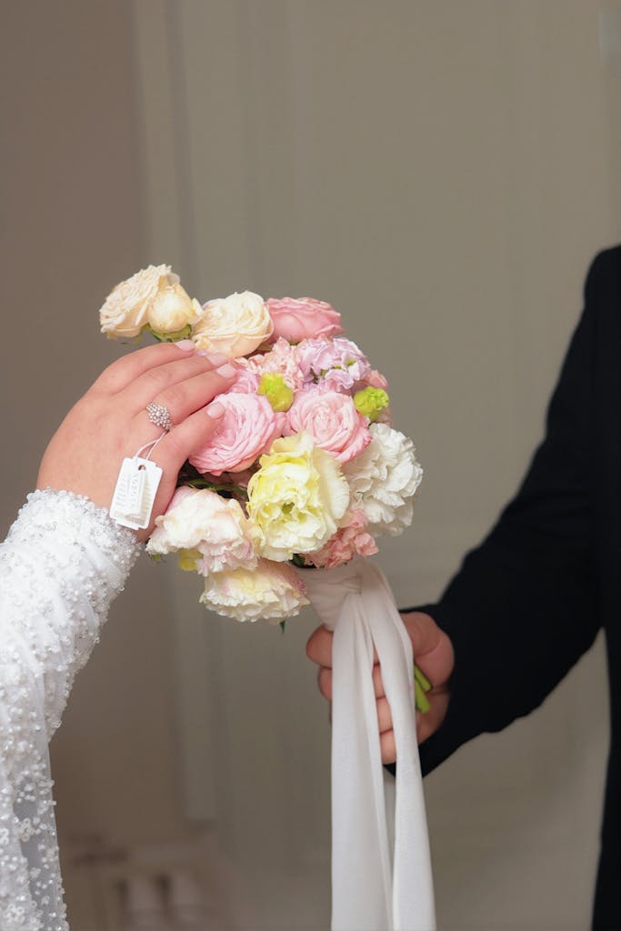 A person in a white, beaded outfit holds a bouquet of pink and white flowers while another in a dark suit grasps the bouquet’s ribbon—a classic moment in wedding photography. The hand in white wears a ring and tag, against a plain, light-colored background. Infinity Photography - Wedding Photographer - East Sussex