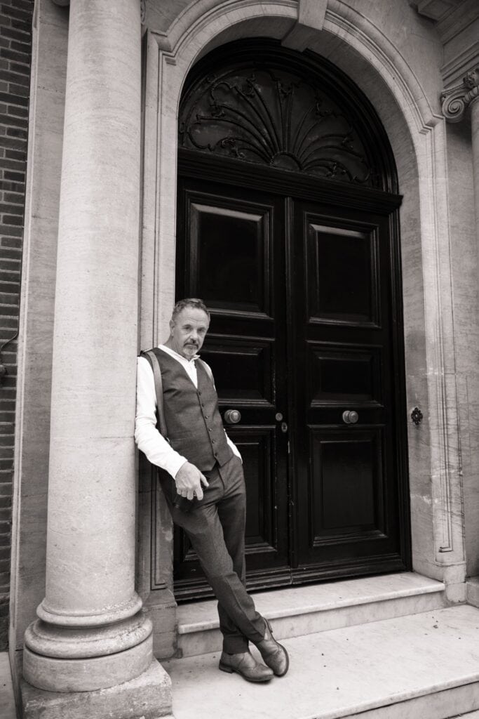 A man in a vest and dress shirt leans casually against a column in front of a tall, ornate black door with decorative arch details—a timeless black-and-white moment that exudes the elegance of classic wedding photography. Infinity Photography - Wedding Photographer - East Sussex